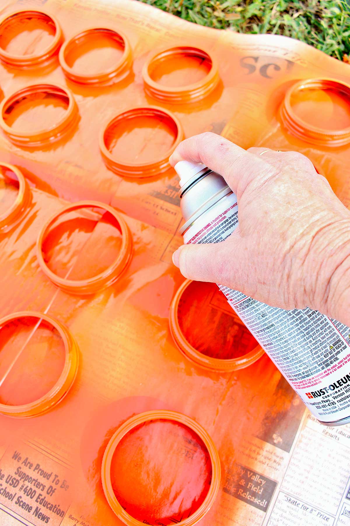 Person spray painting mason jar lids orange outside on a piece of newsprint.