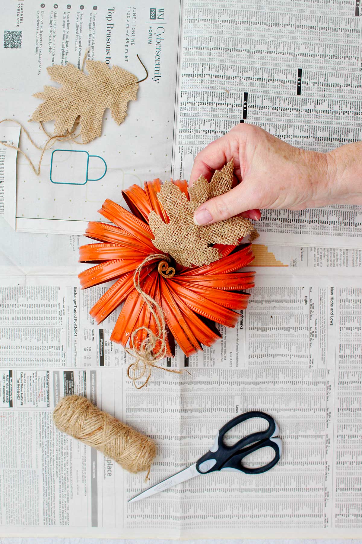 Person attaching a leaf cut out from burlap on the top of the mason jar lid pumpkin.