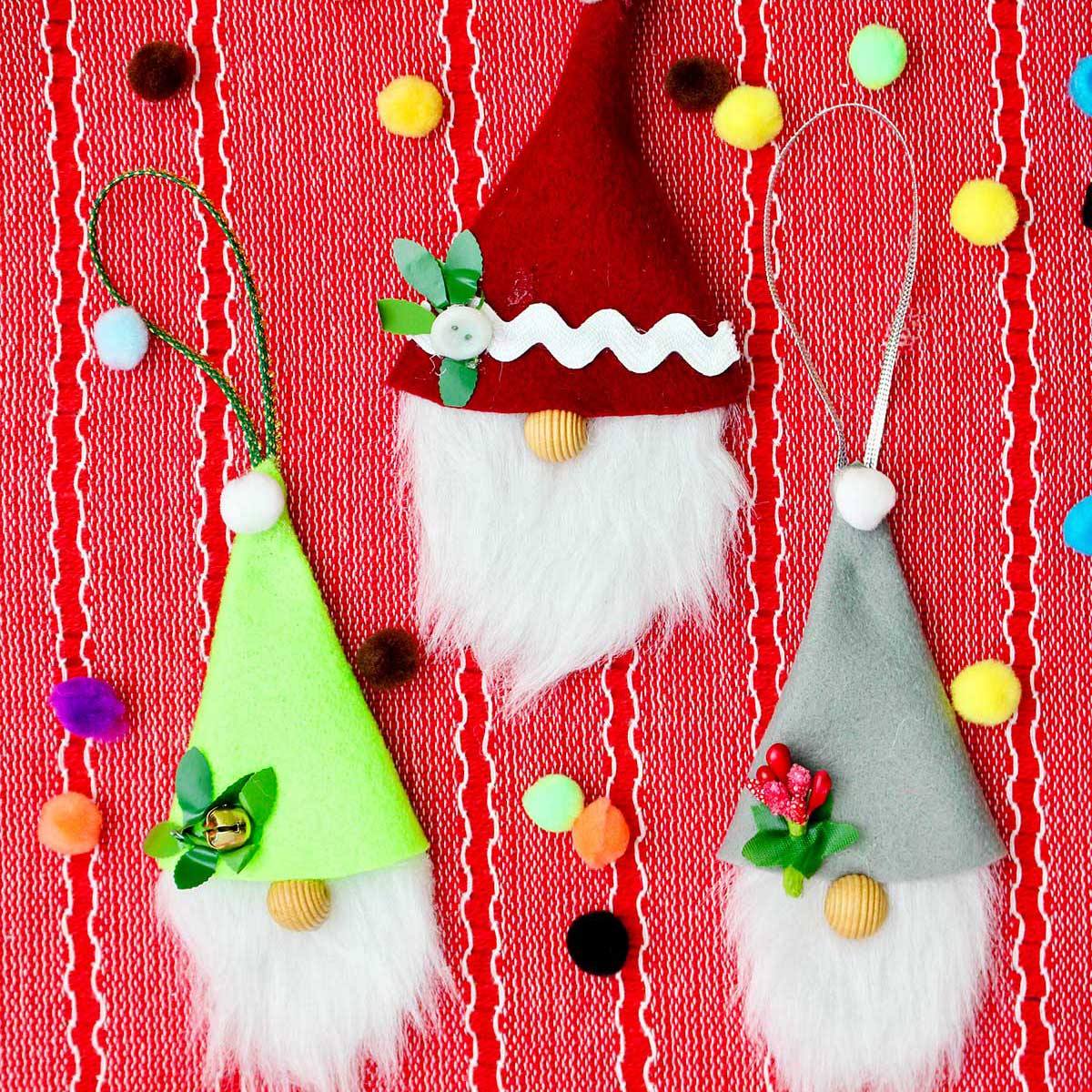 Three completed gnome Christmas ornaments on a red placemat with colorful pom poms scattered around.