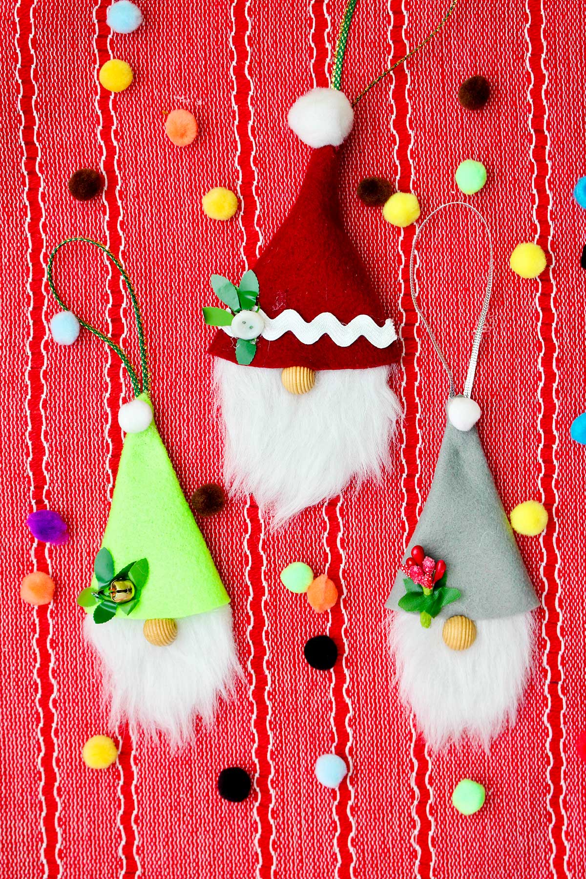 Three completed gnome Christmas ornaments on a red placemat with colorful pom poms scattered around.