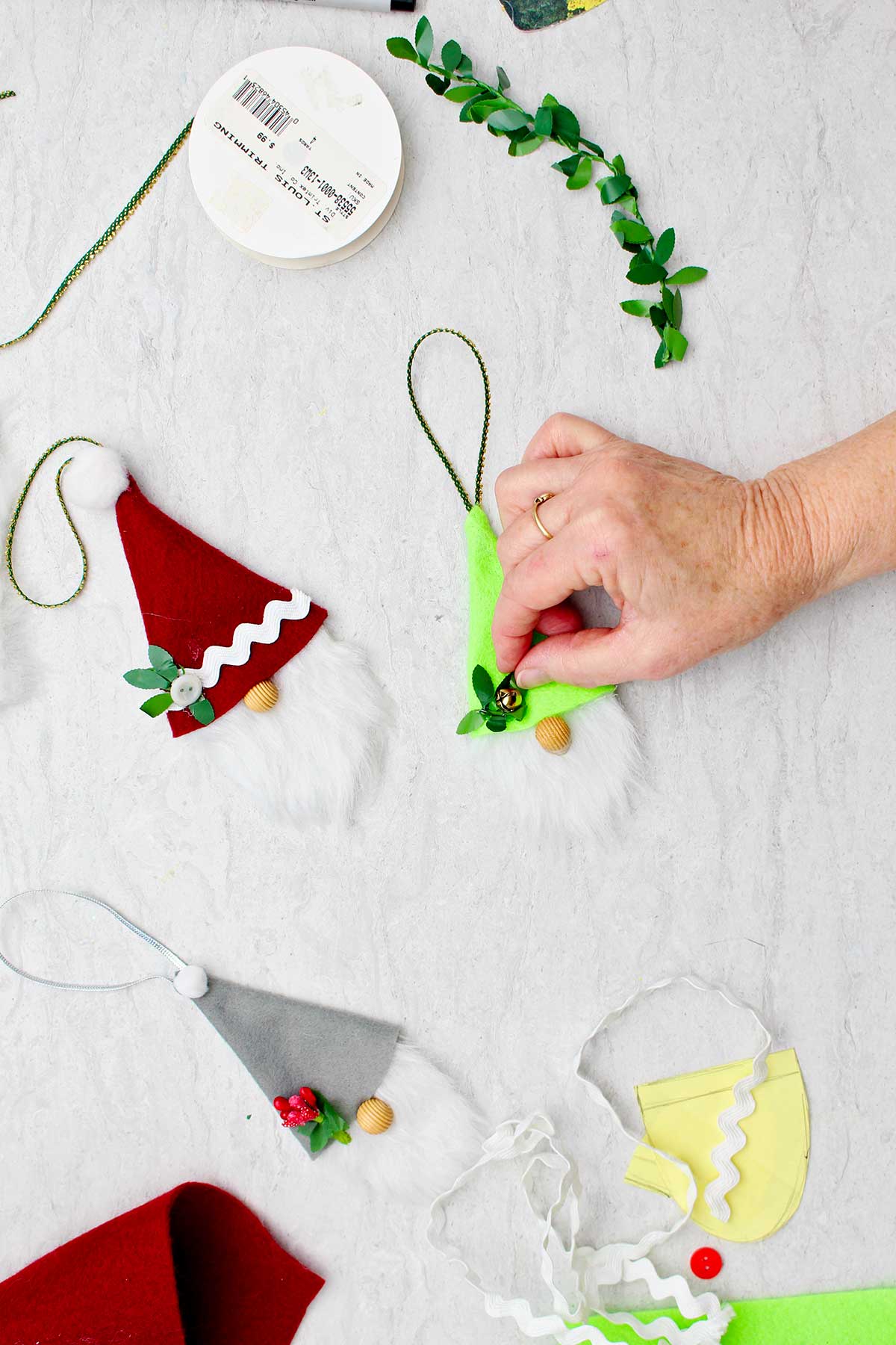 Person adding a bell as a decoration to a green hat on a gnome ornament with other completed gnome ornaments and supplies near by.