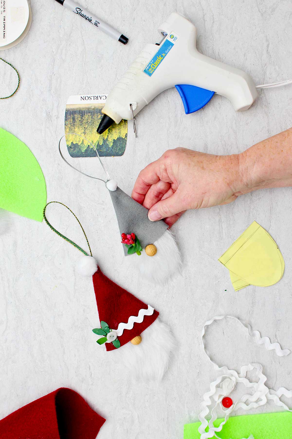 Person holding a completed gnome Christmas ornament wearing a grey felt hat with another completed ornament and supplies near by.