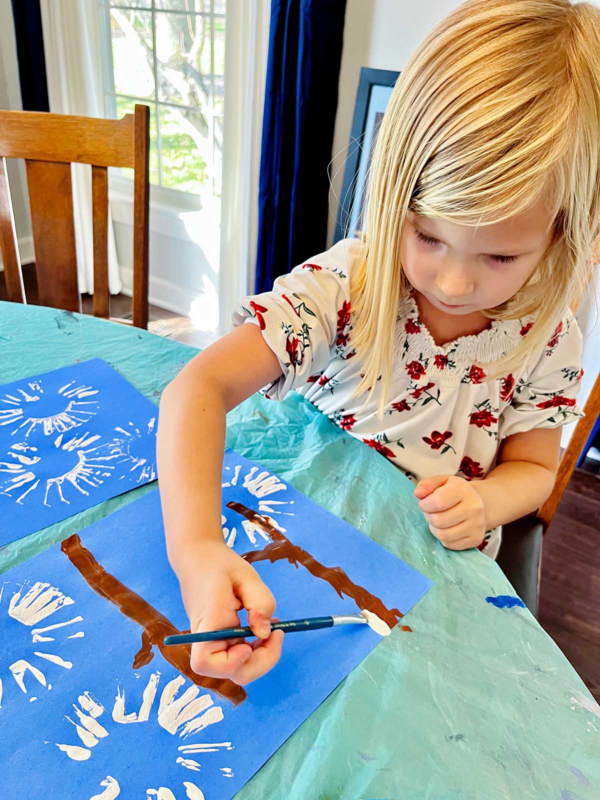 Young blonde girl with floral shirt adds white paint for snow on branches in her cute winter cardinal handprint craft.