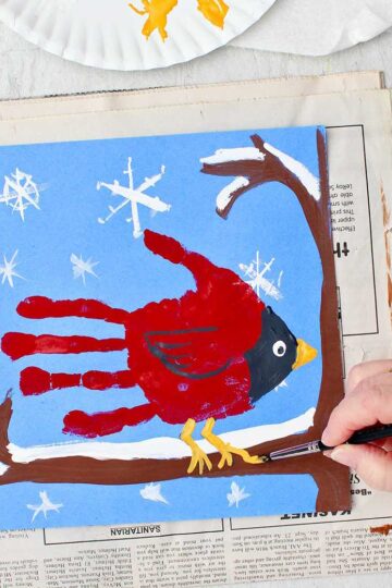 Person painting final details on a cute winter cardinal handprint painting of a cardinal handprint sitting on a snowy branch.