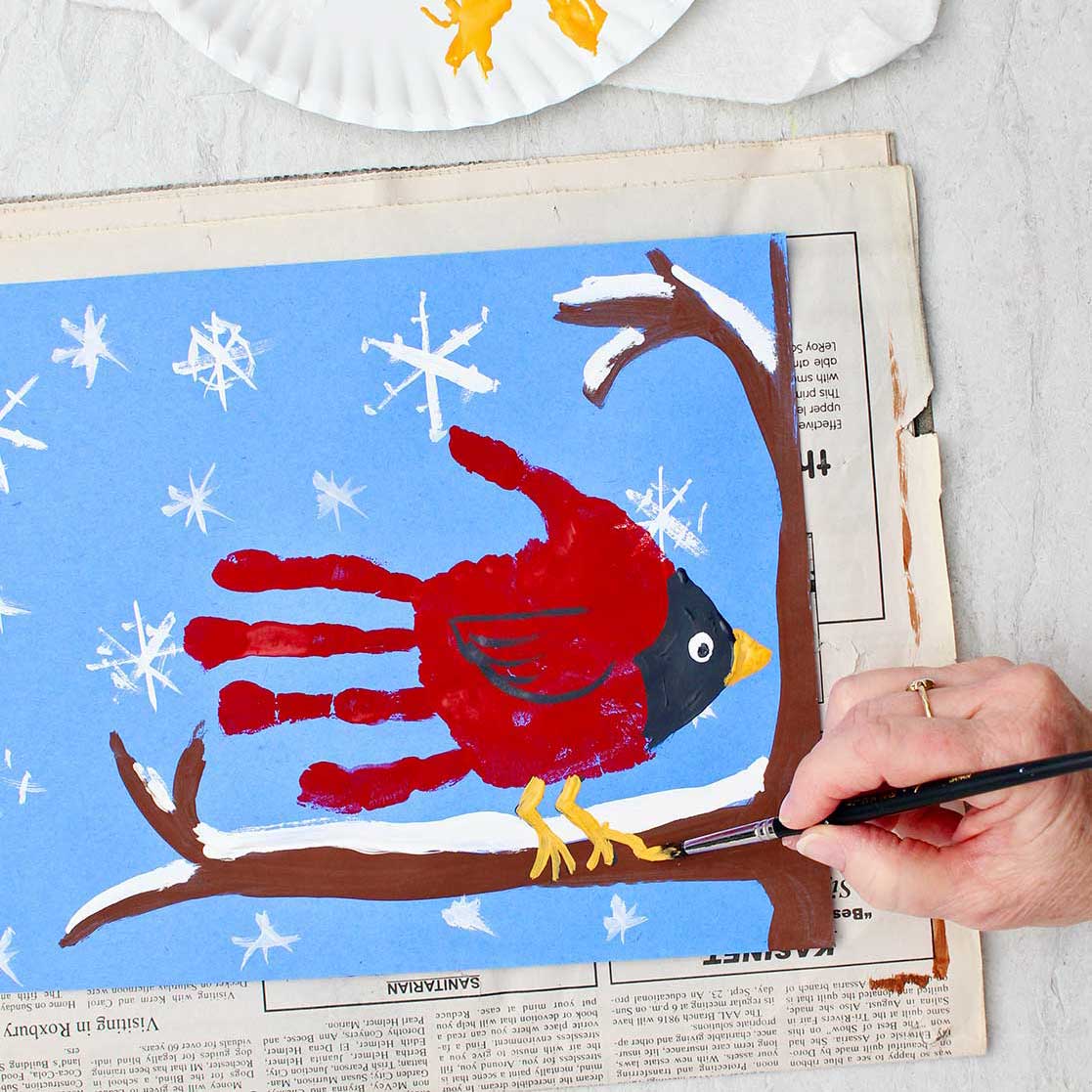 Person painting final details on a cute winter cardinal handprint painting of a cardinal handprint sitting on a snowy branch.