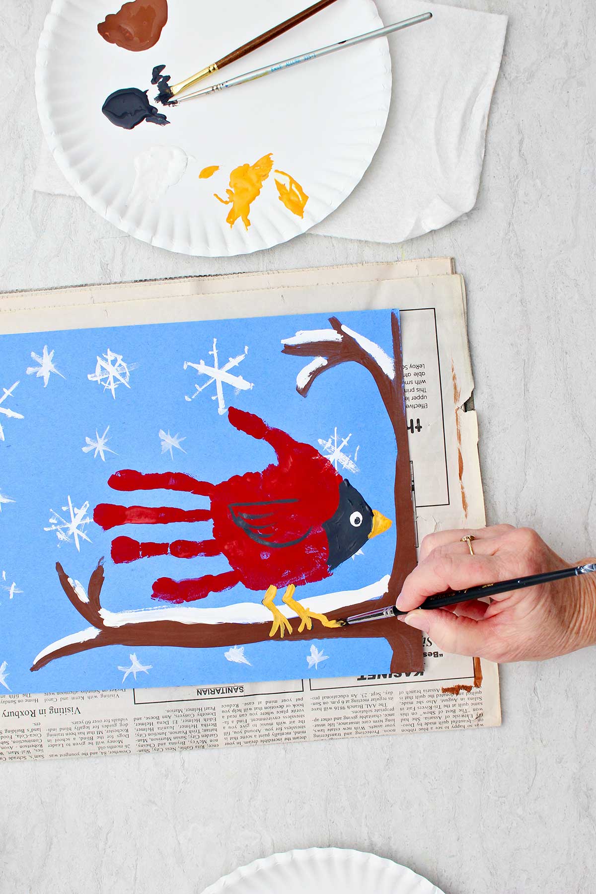 Person painting final details on a cute winter cardinal handprint painting of a cardinal handprint sitting on a snowy branch.