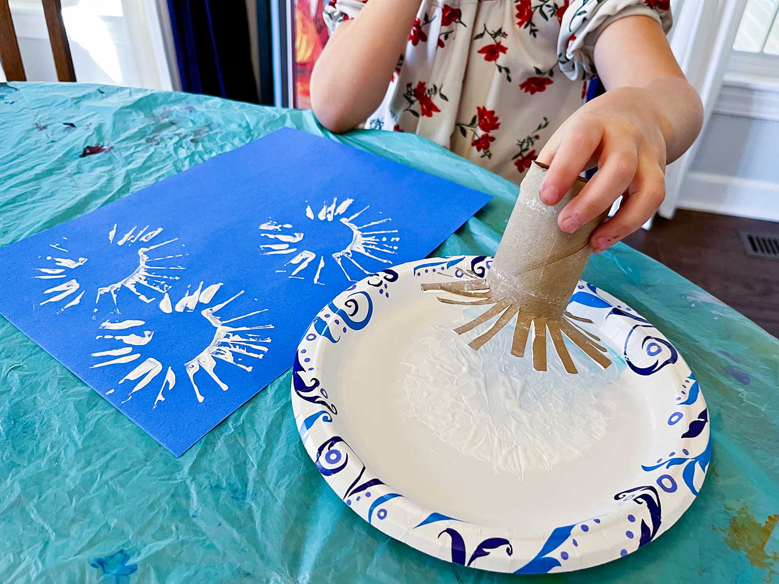 Young girl using a toilet paper towel roll cut like a sunburst to make stamps for her project on a blue piece of construction paper.