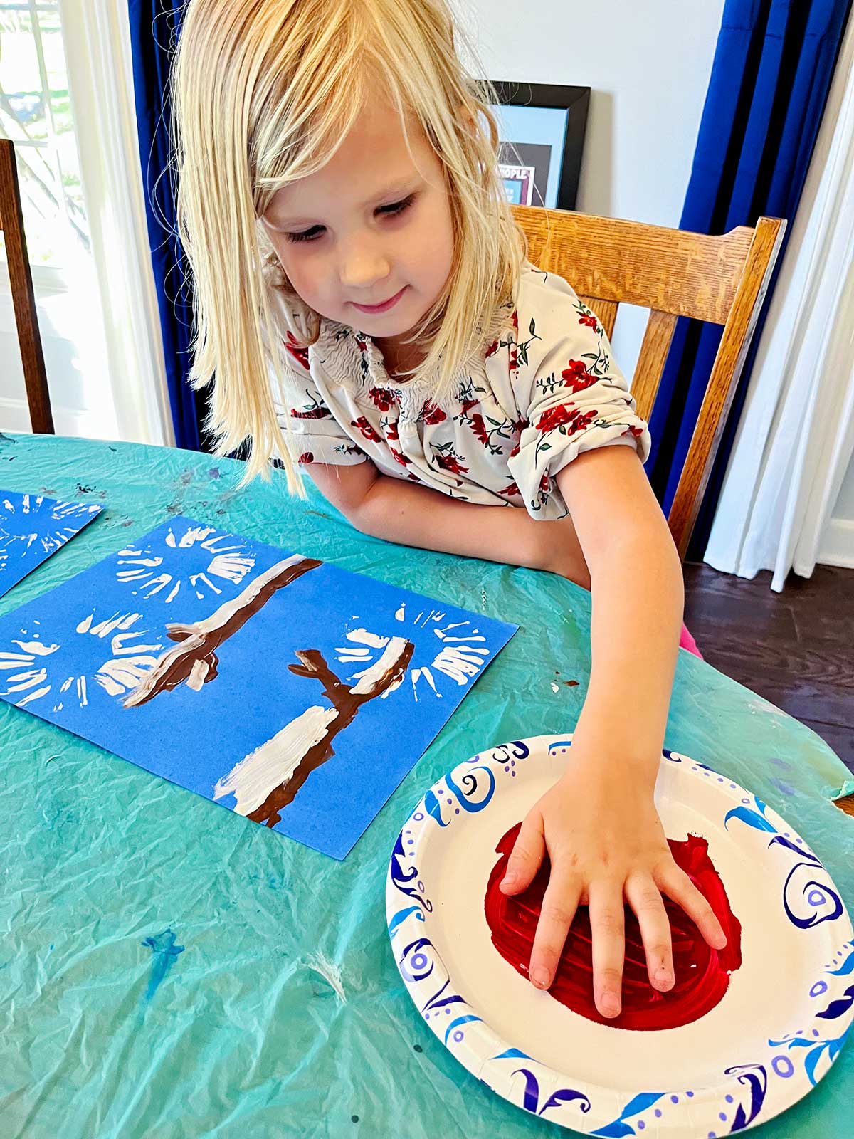 Young blonde girl with floral shirt puts her hand in red paint for her cute winter cardinal handprint craft.