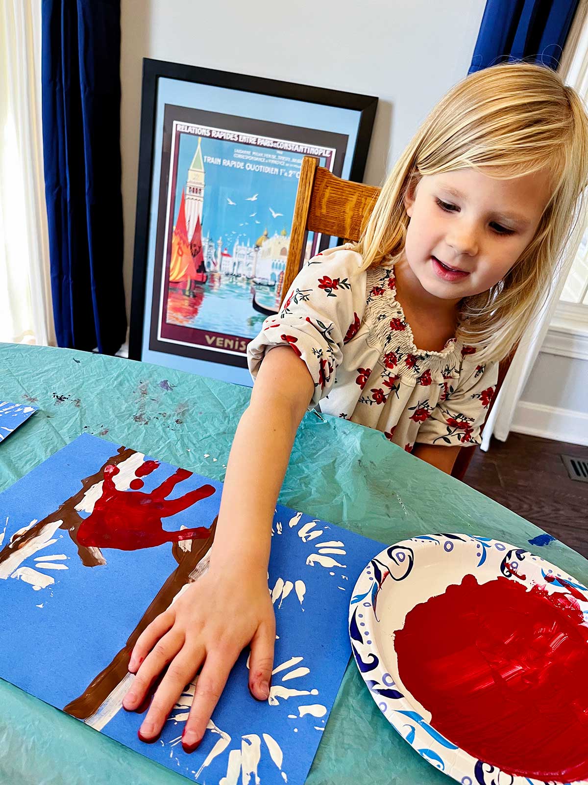 Young blonde girl with floral shirt puts a red handprint on her cute winter cardinal handprint craft.