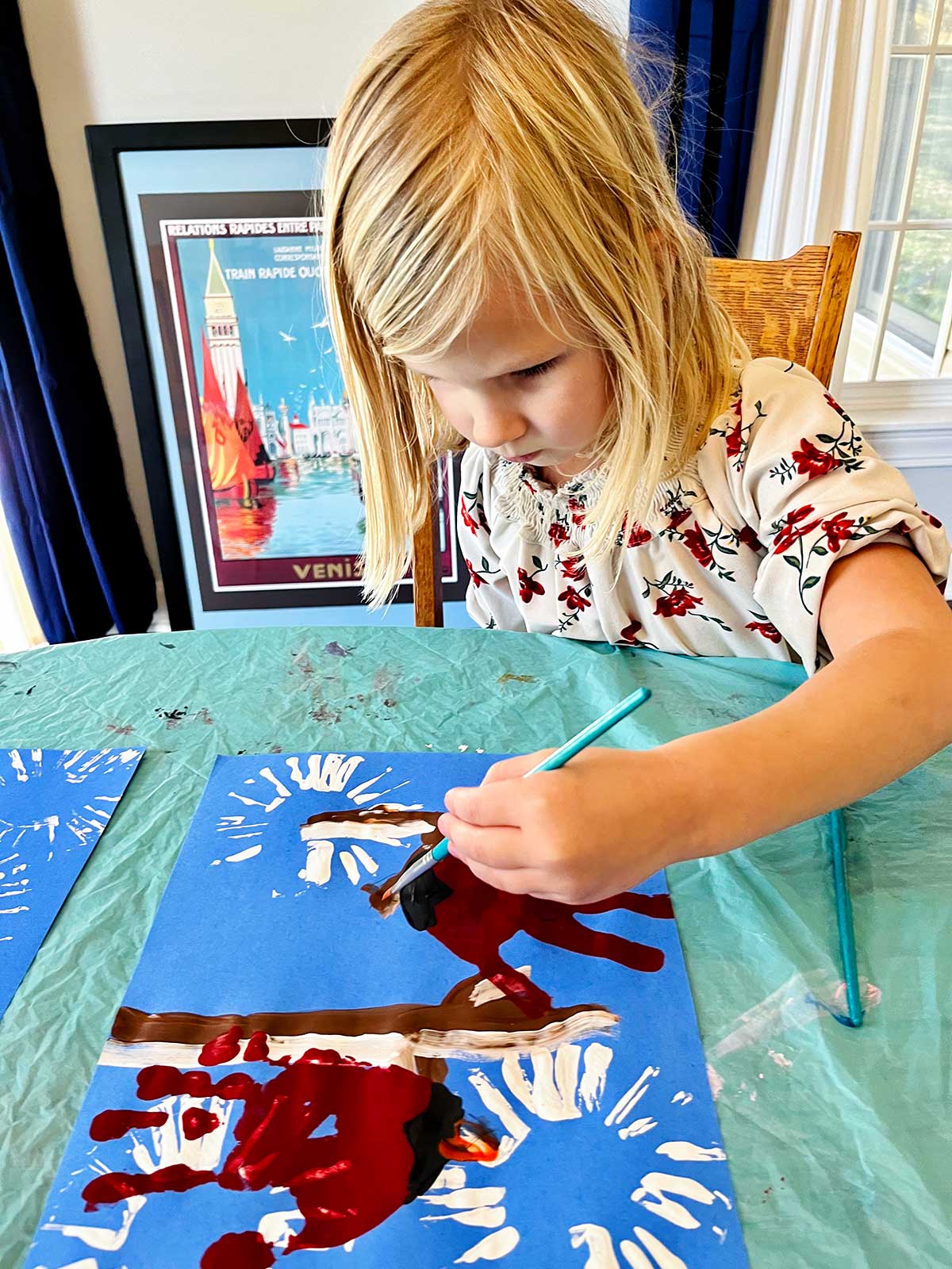 Young blonde girl with floral shirt puts details on her cute winter cardinal handprint craft.