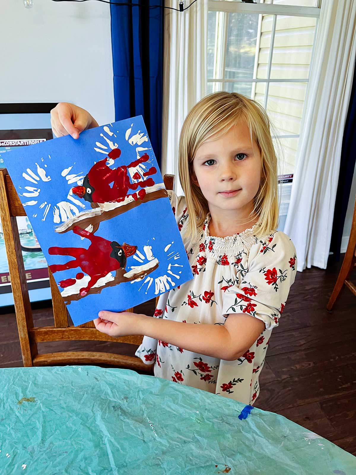 Young blonde girl with floral shirt holds up her completed cute winter cardinal handprint craft.