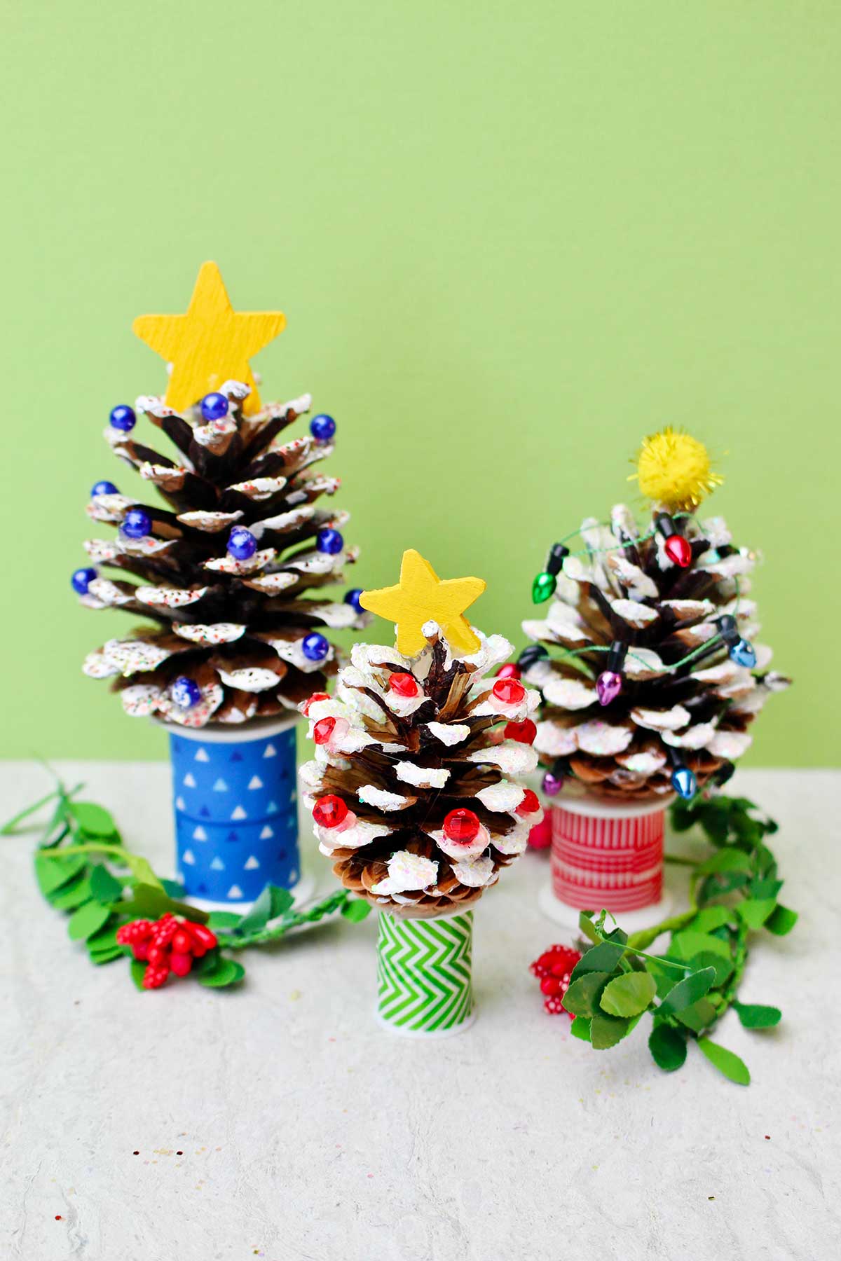 Three pinecones painted with white "snow", decorated with beads and Christmas lights, glitter, and a painted yellow star to look like Christmas trees