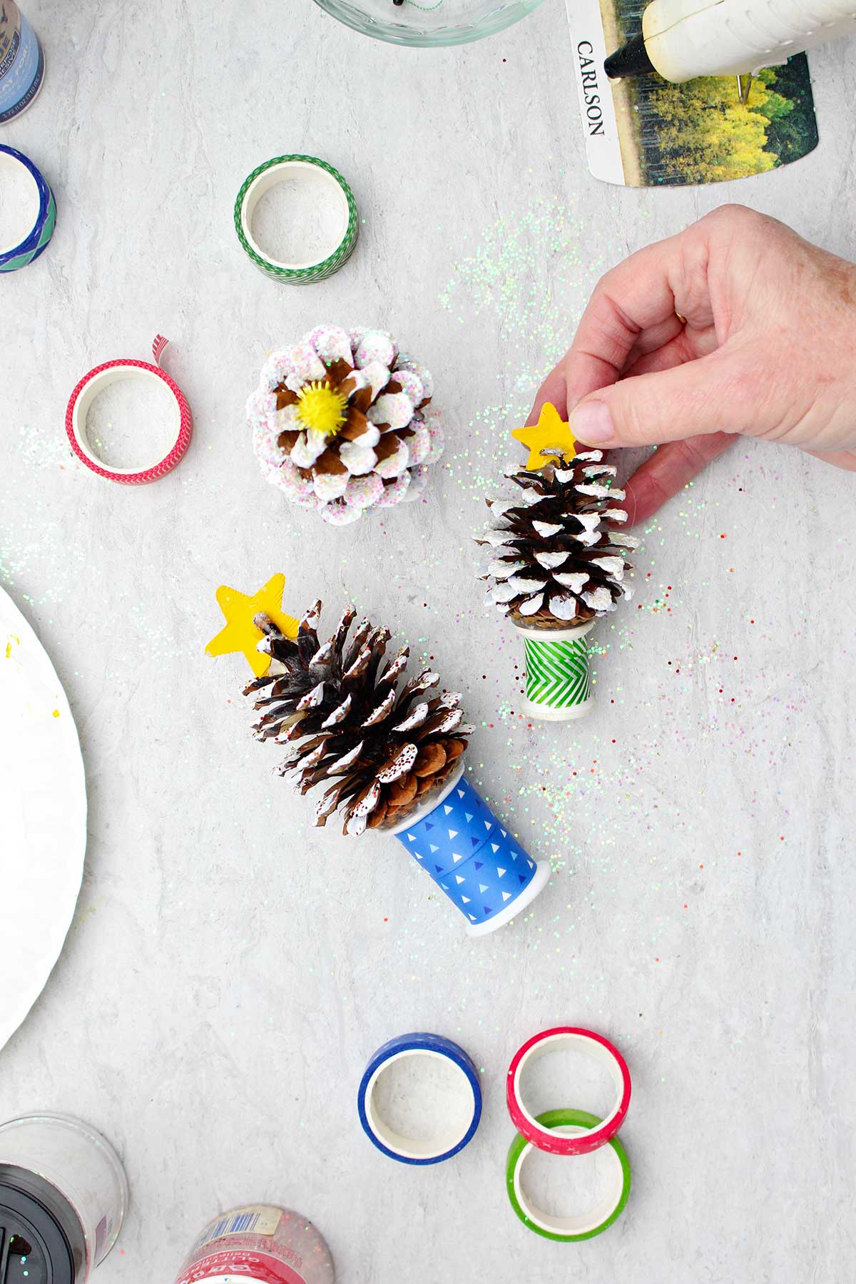 Adding a painted yellow star to a frosted pinecone Christmas tree near ribbons, glitter, and a hot glue gun