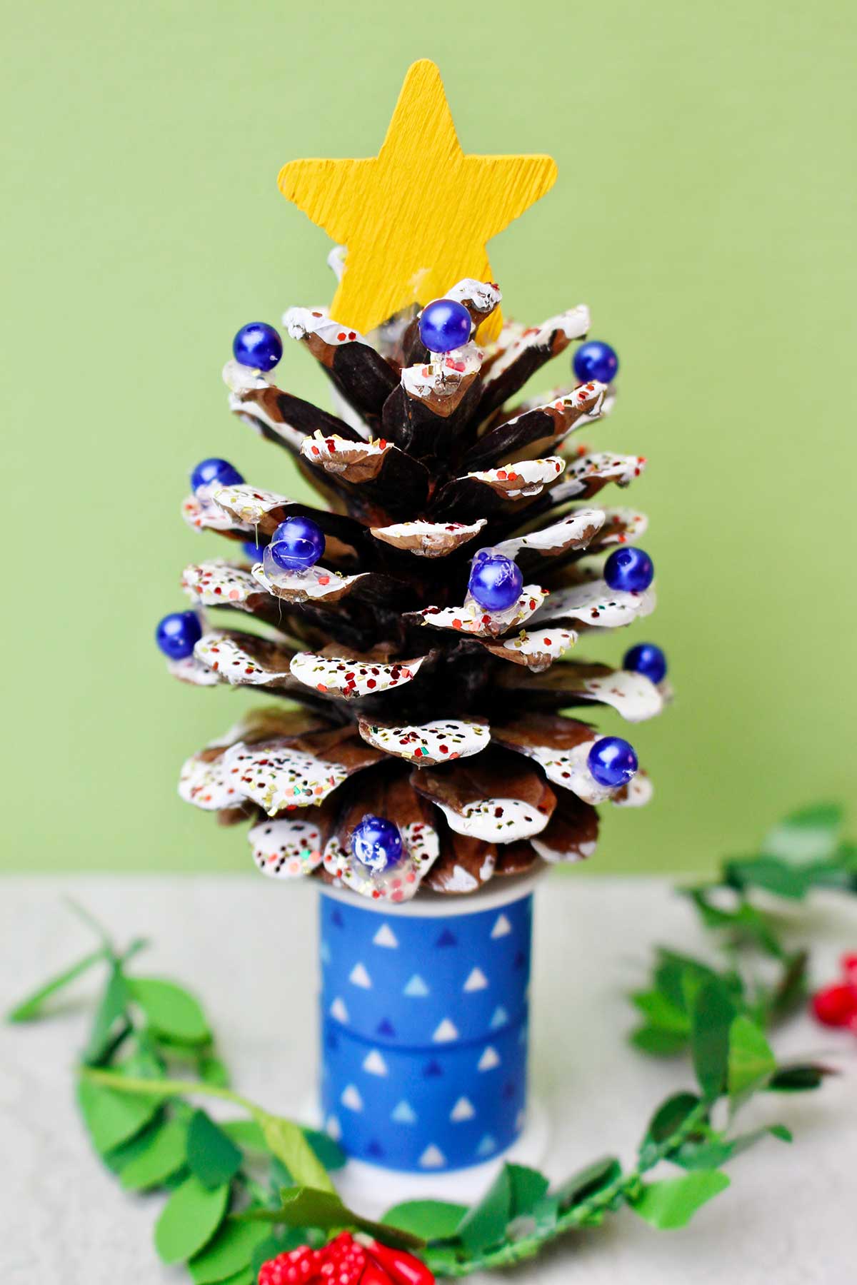 A pinecone painted with white "snow", decorated with blue beads, glitter, and a yellow painted star to look like a Christmas tree