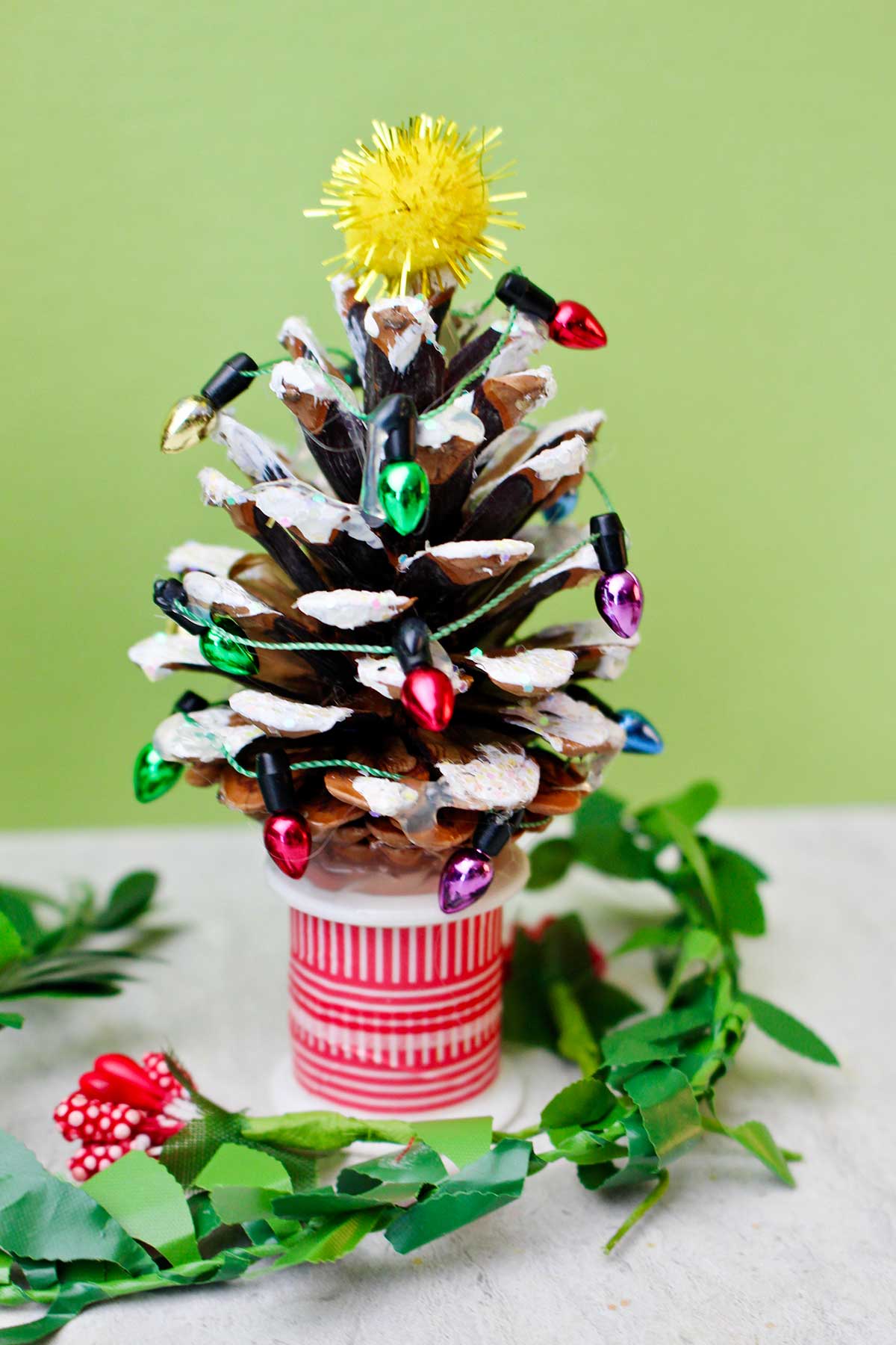 A pinecone painted with white "snow", decorated with mini Christmas lights, glitter, and a yellow painted star to look like a Christmas tree