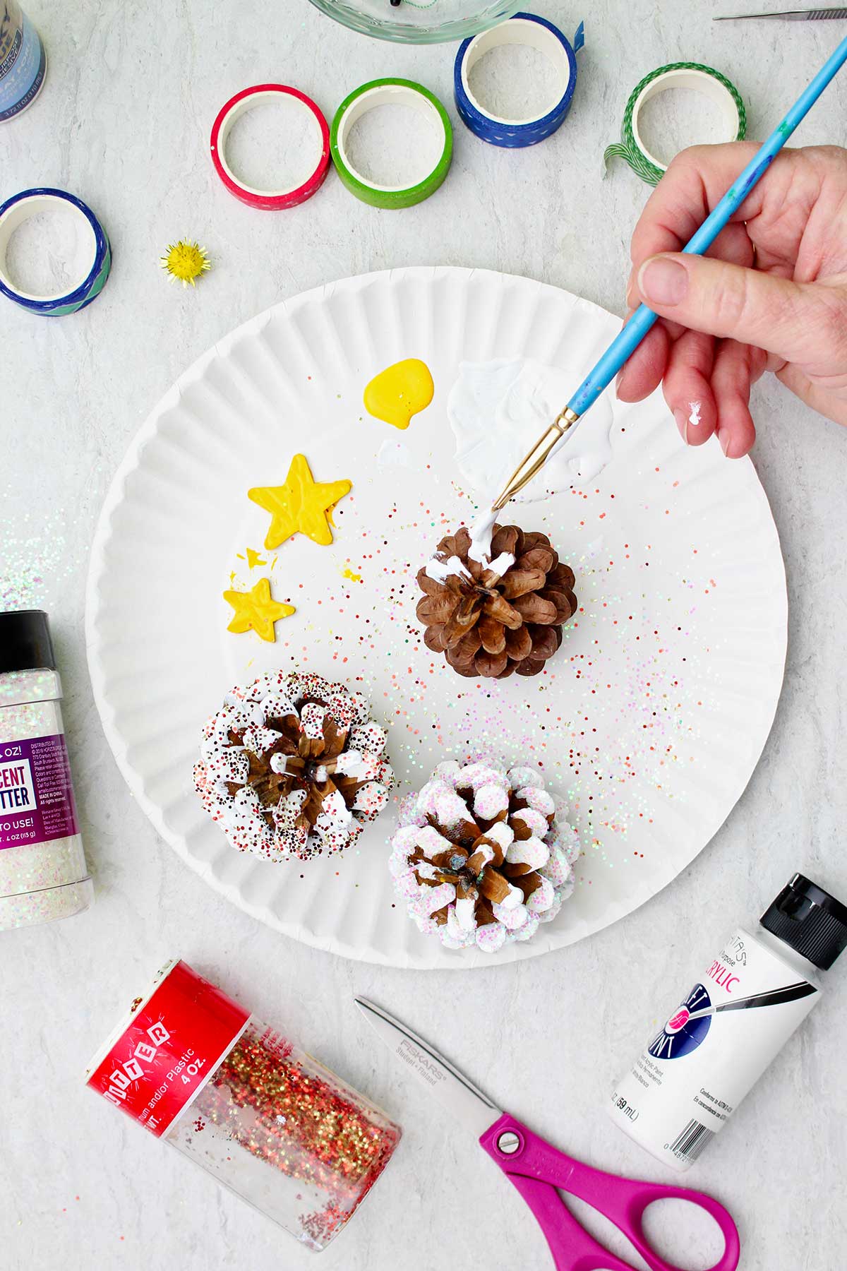 White painted being painted on the edges of a pinecone like "snow", near painted yellow stars