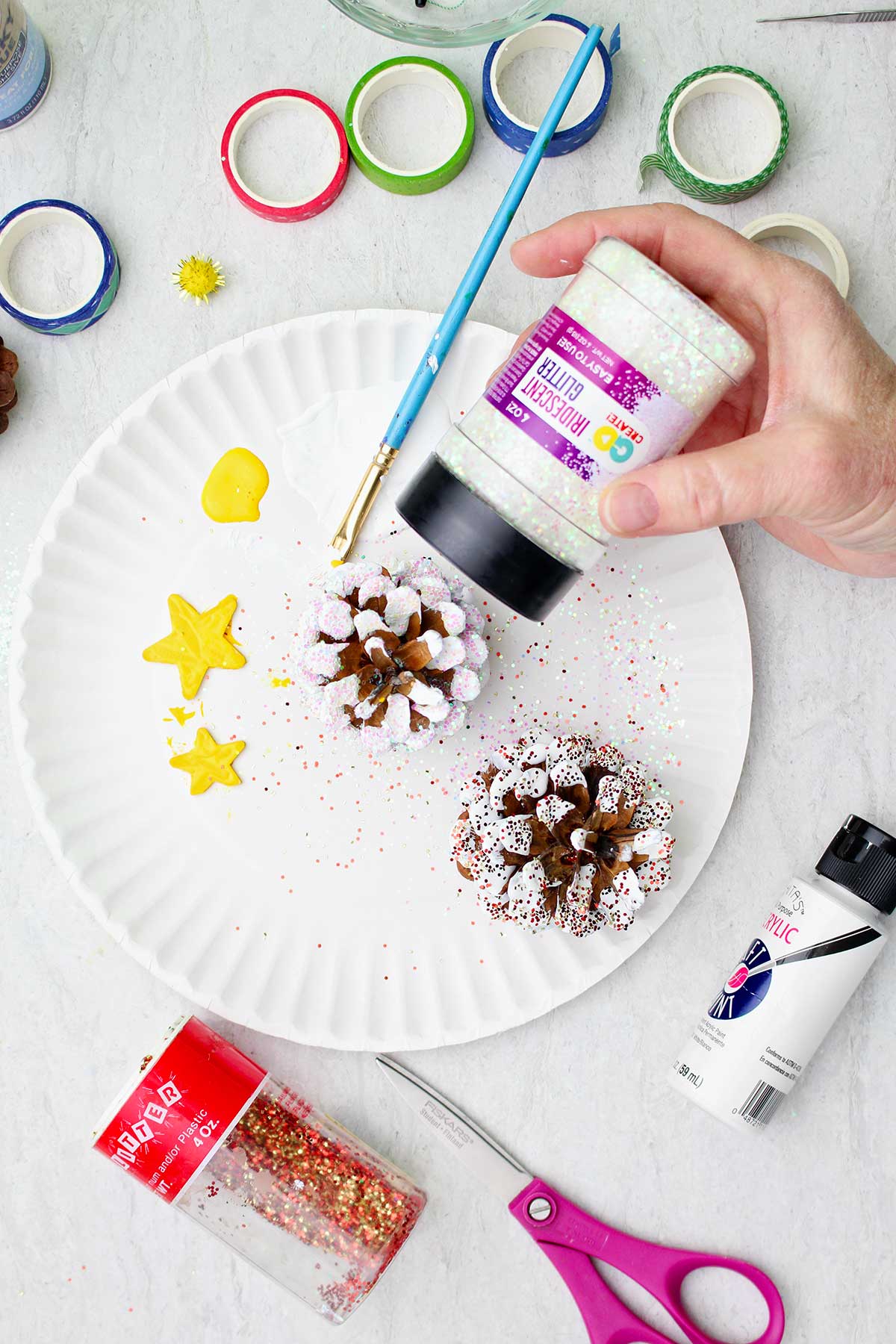 Glitter being sprinkled onto a pinecone, painted with white "snow"