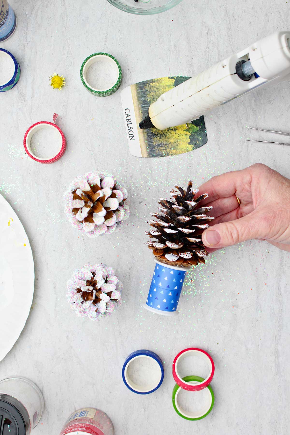 A hand gluing a frosted pinecone Christmas tree to a spool covered with blue ribbon