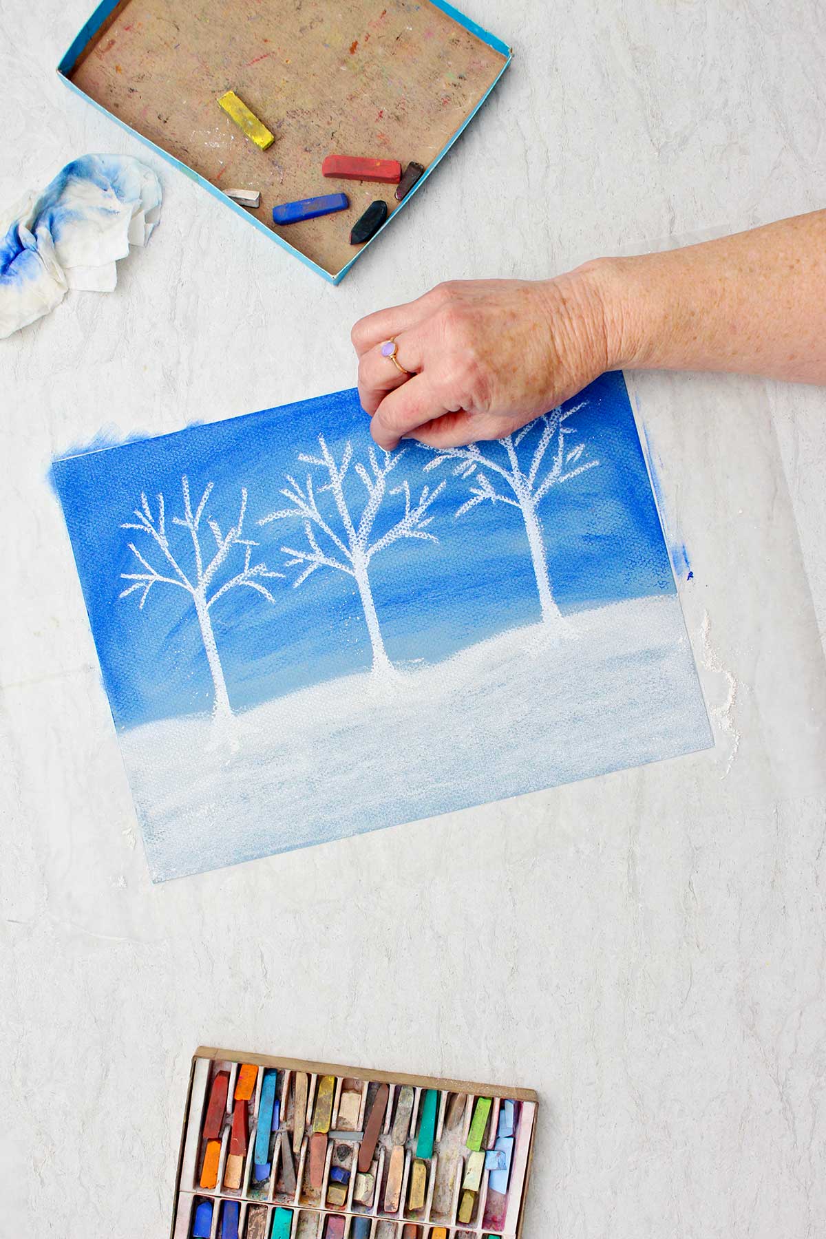 Person adding branch details to white trees on pastel chalk drawing with a box of pastels near by.