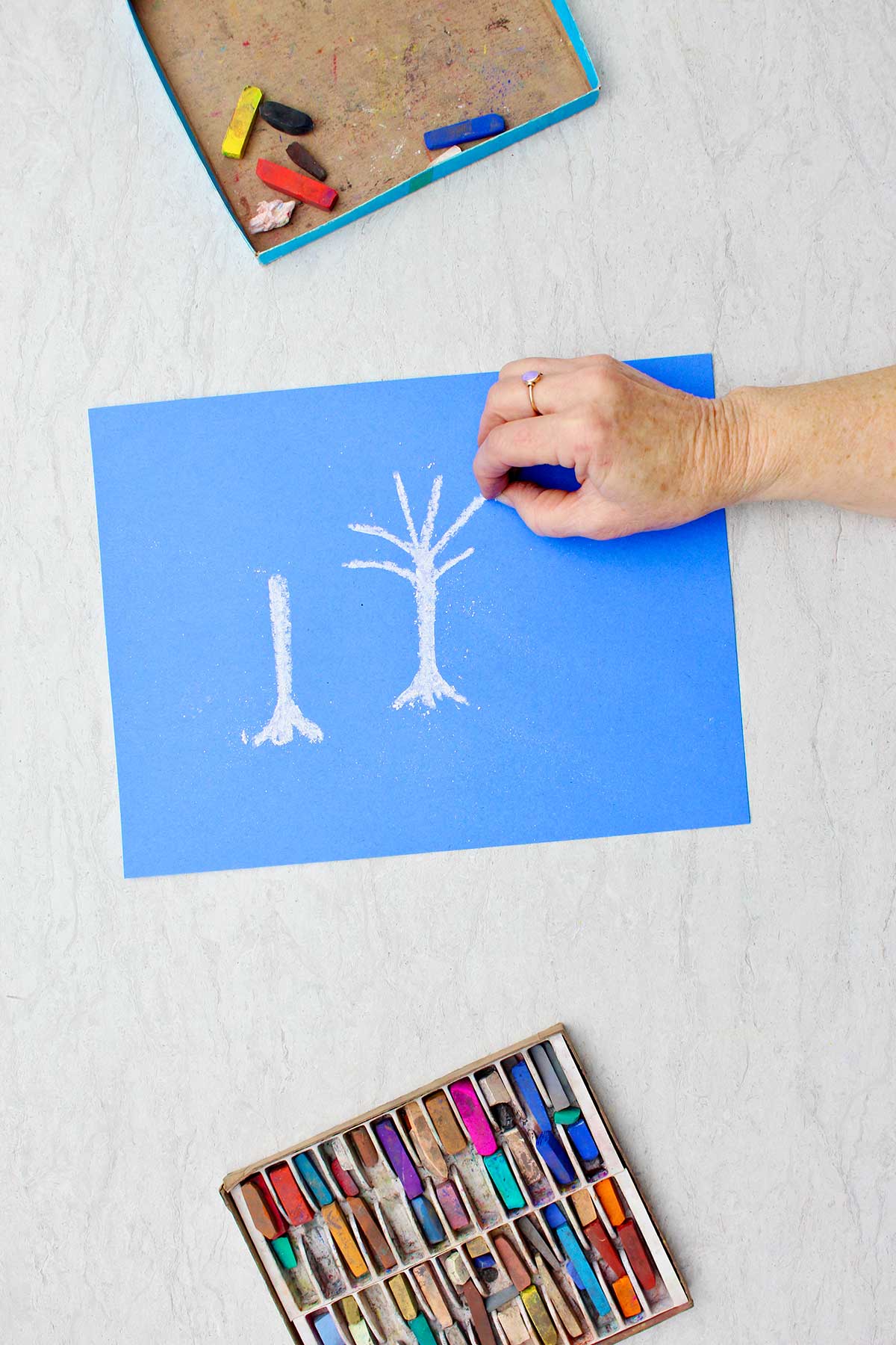 Person drawing white trees with pastel chalk on a blue piece of paper with a box of pastels near by.