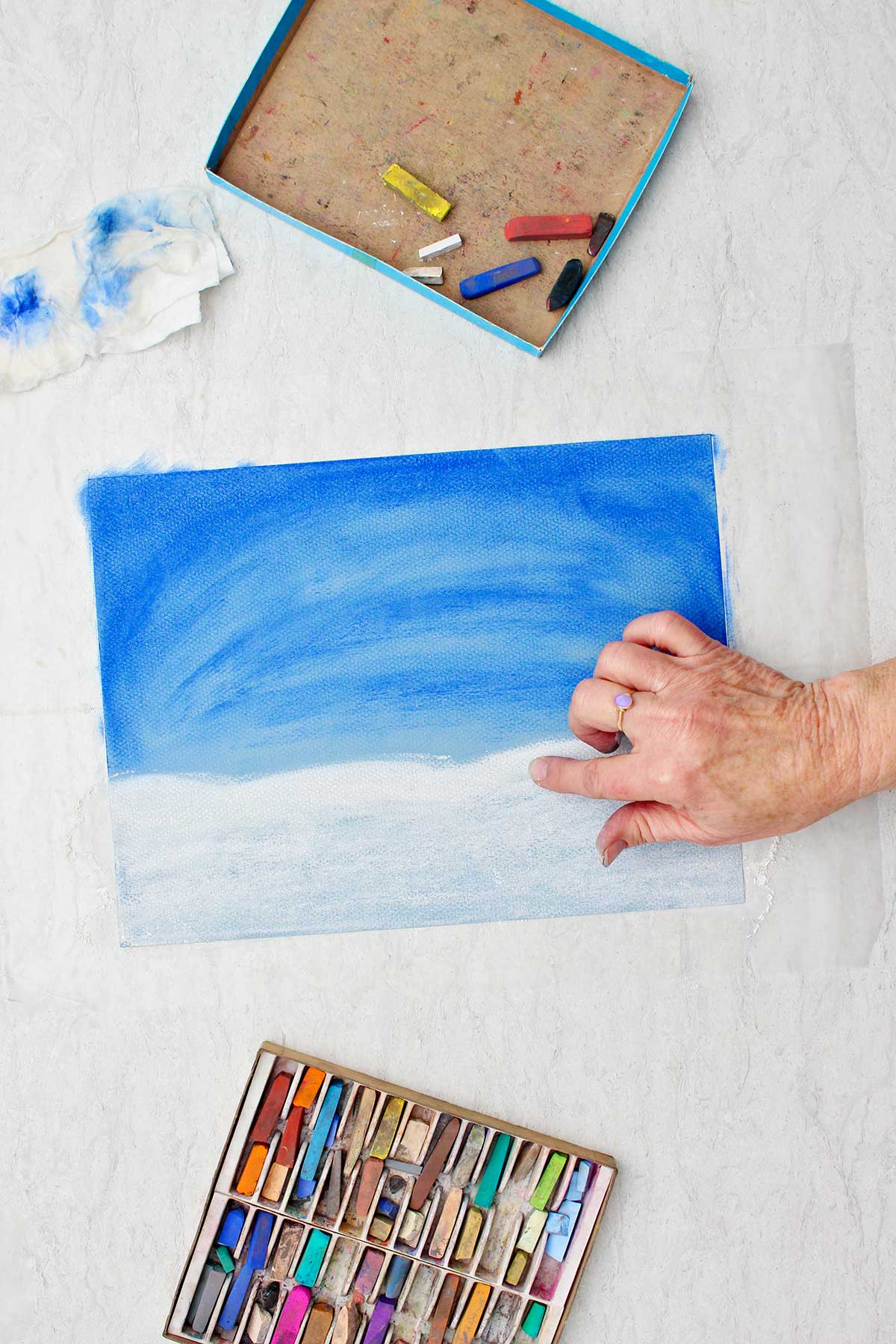 Person blending some white pastel chalk on a paper to look like snow with a box of pastel chalk near by.