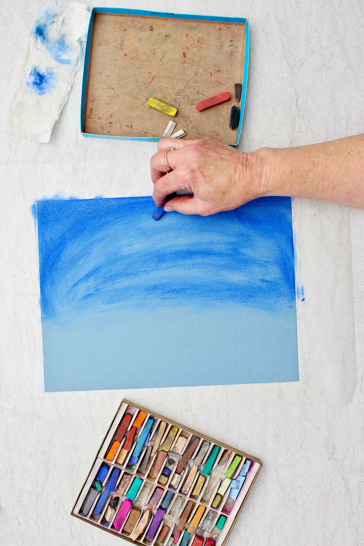 Person using a piece of blue pastel chalk to darken up the sky on a blue piece of paper with a box of pastel chalks near by.