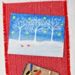 A winter scene of white trees and show against a blue night sky with stars on paper colored with pastels sitting on a red placemat with pieces of pastels near by.