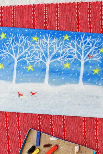 A winter scene of white trees and show against a blue night sky with stars on paper colored with pastels sitting on a red placemat with pieces of pastels near by.