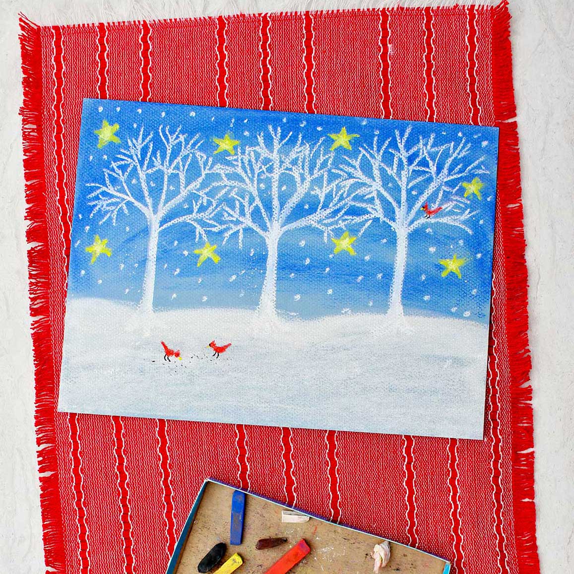 A winter scene of white trees and show against a blue night sky with stars on paper colored with pastels sitting on a red placemat with pieces of pastels near by.