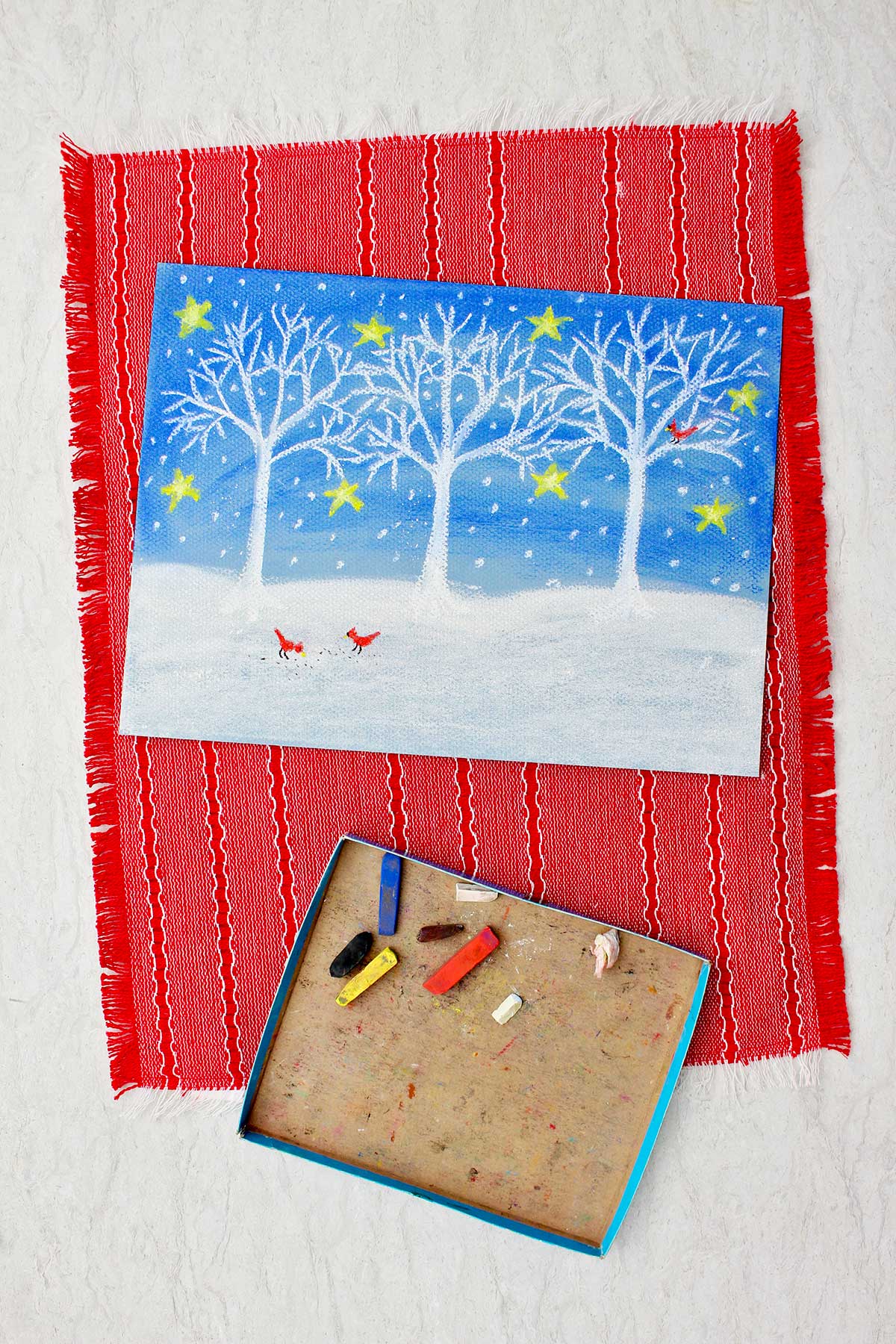 A winter scene of white trees and show against a blue night sky with stars on paper colored with pastels sitting on a red placemat with pieces of pastels near by.