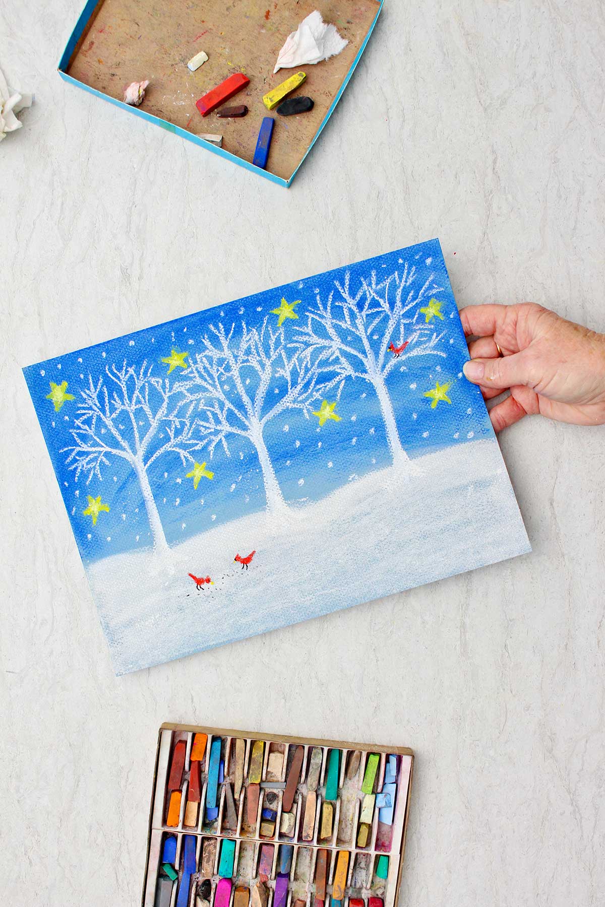 Person holding completed Christmas winter scene colored with pastels with a box of pastel chalk near by.