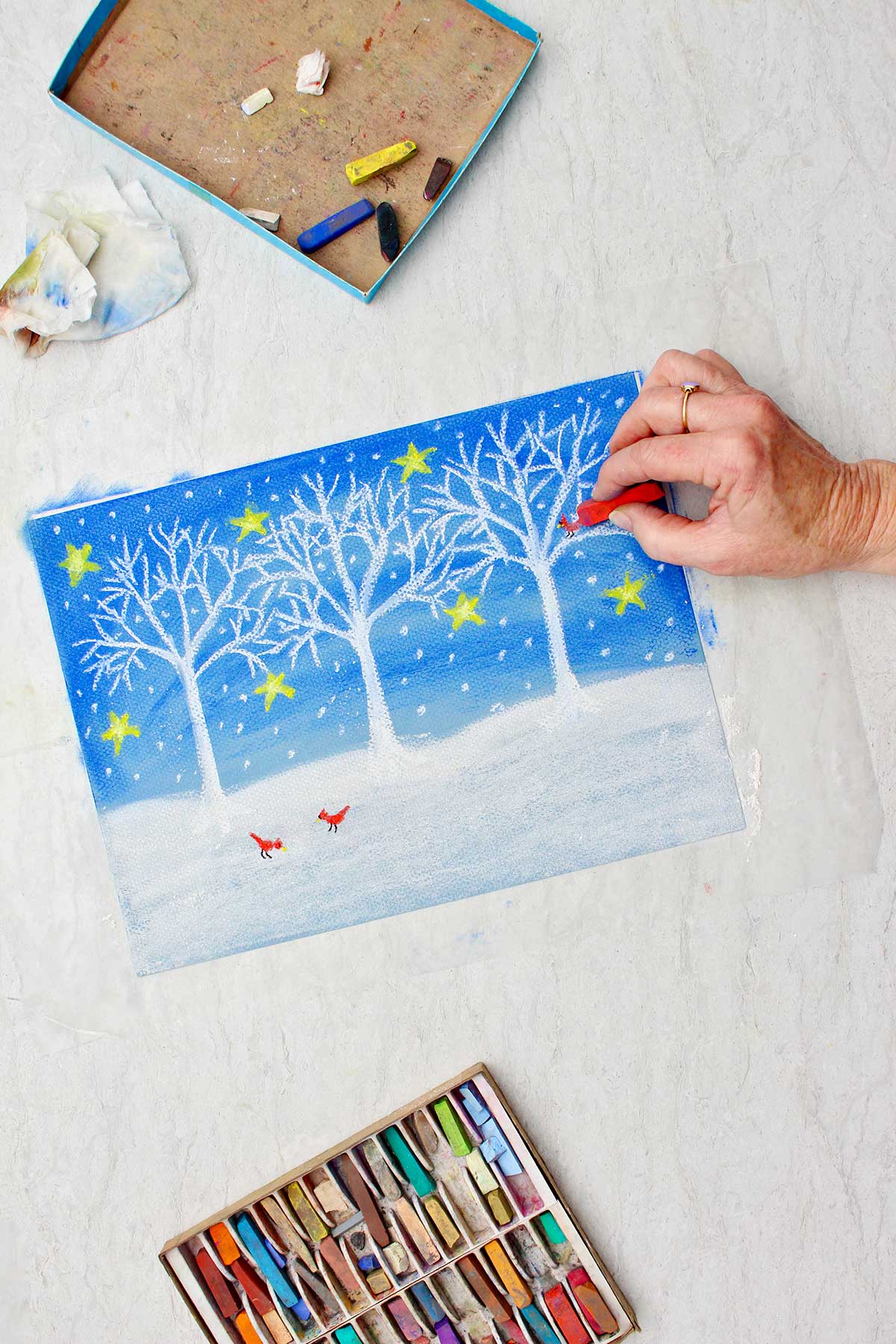 Person adding red cardinals in a tree on an almost completed Christmas winter scene drawing with a box of pastel chalk near by.