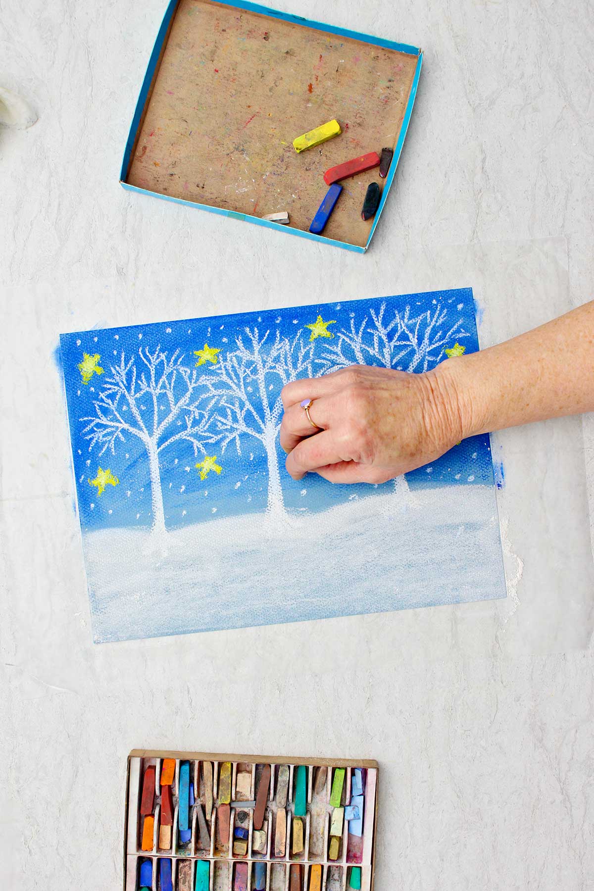 Person adding small white dots to look like snow on Christmas winter pastel drawing with a box of pastels near by.