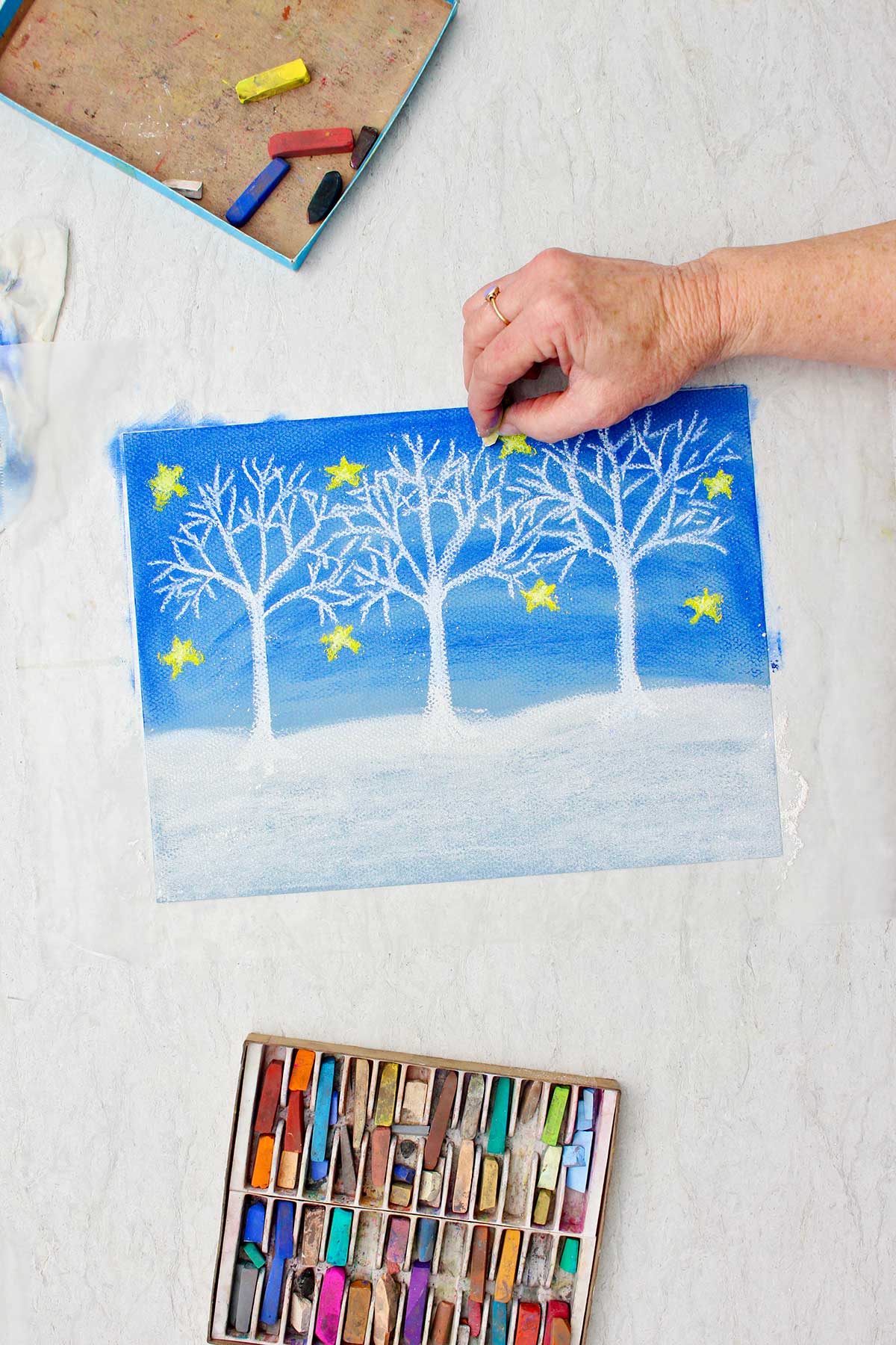 Person adding branch details on a white tree in a Christmas winter drawing with pastel chalk with a box of pastel chalk near by.