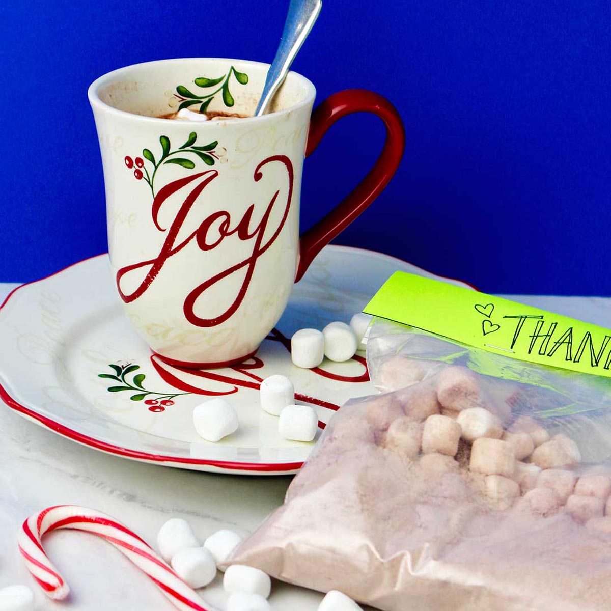 A Joy mug of hot chocolate with marshmallows beside a bag of homemade cocoa mix with a green thank you label and a candy cane