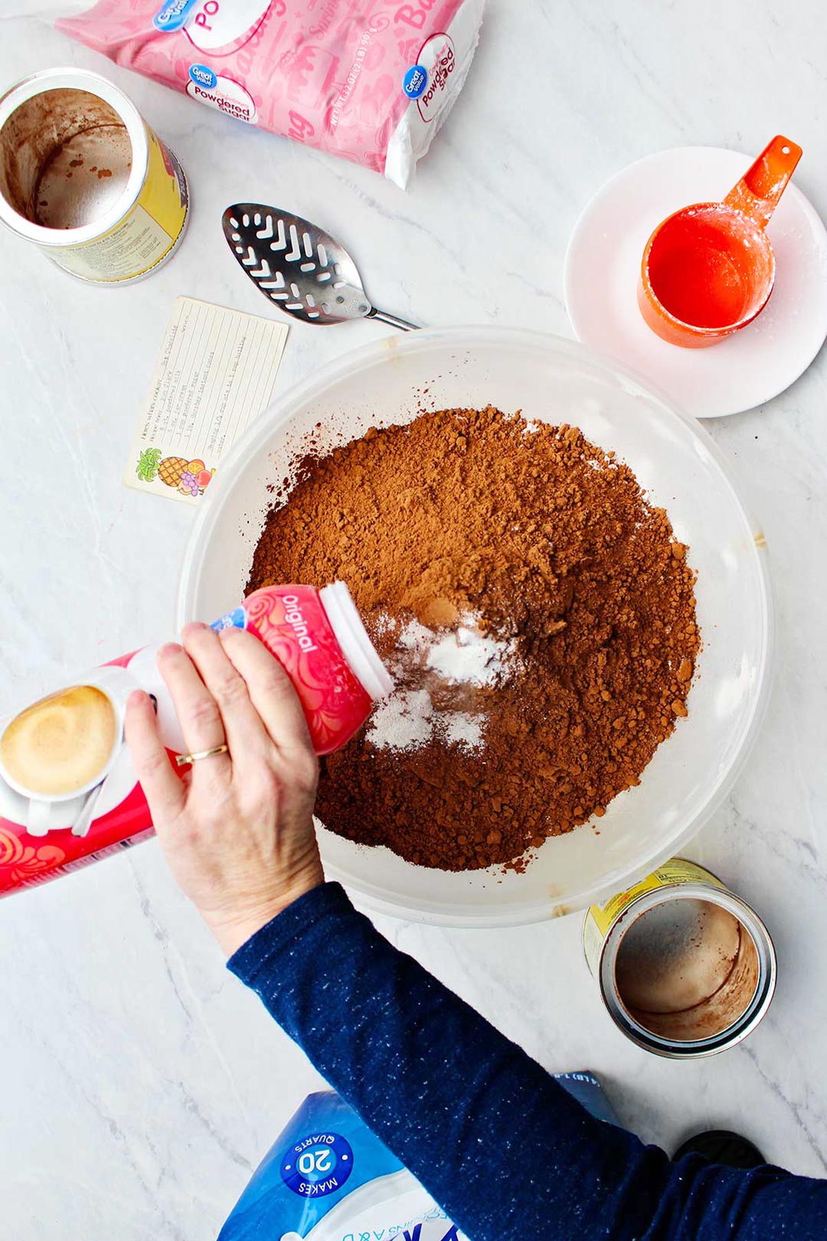 Hand putting powdered creamer in bowl of hot chocolate mix with supplies around on counter.