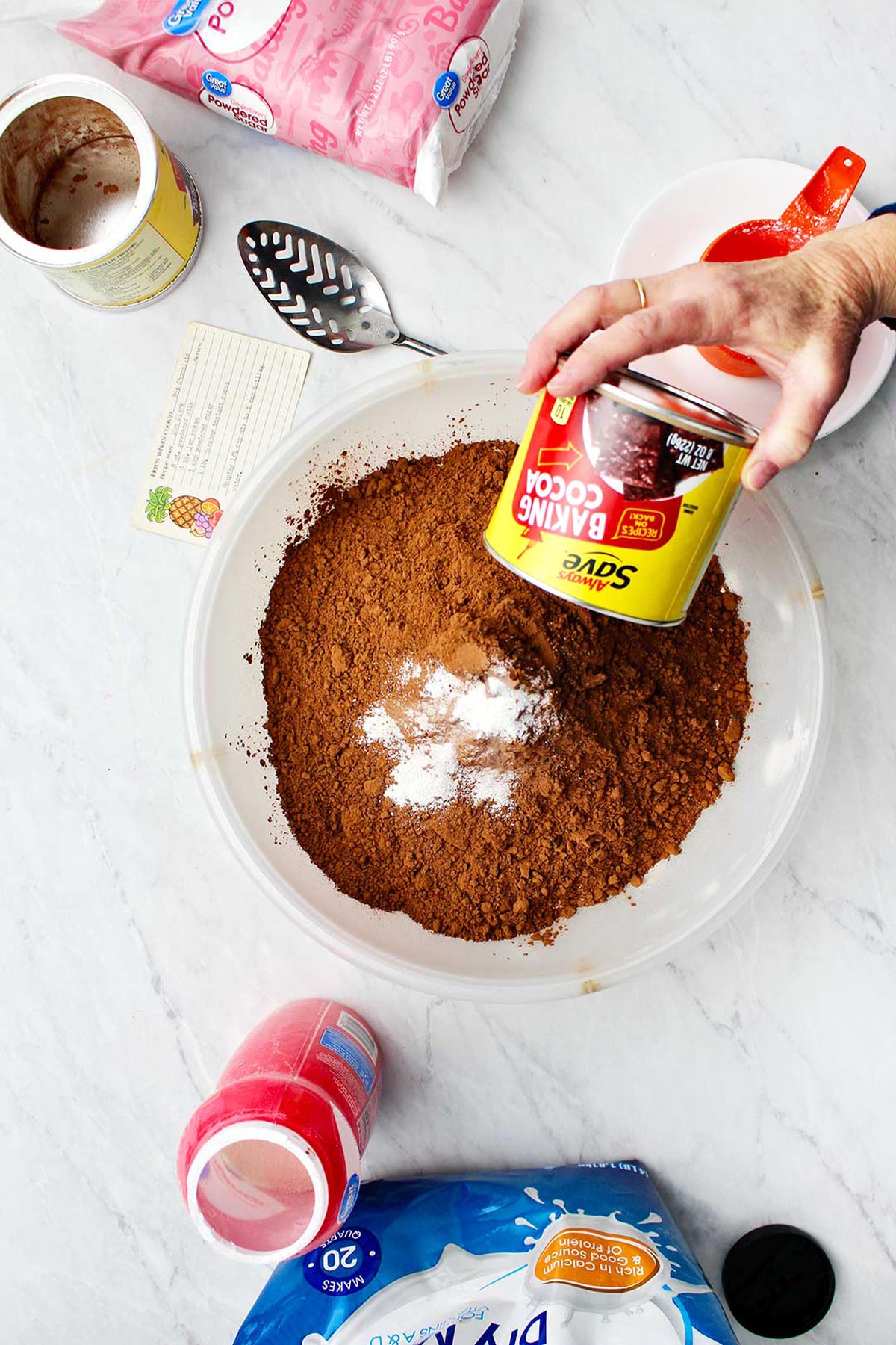 Hand dumping baking chocolate into bowl of hot chocolate mix with supplies near by.