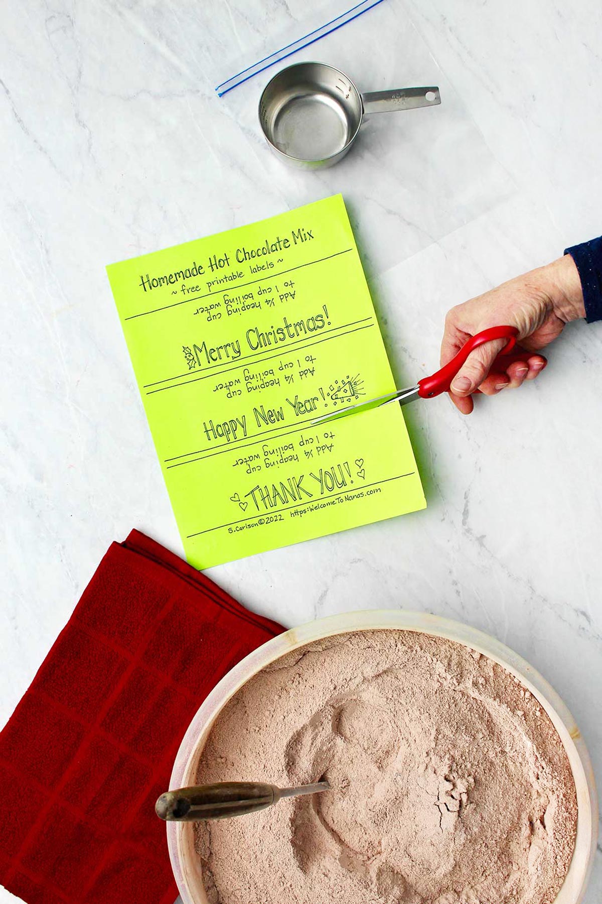 Hand cutting neon green labels for bags of hot chocolate with bowl of hot chocolate mix and red towel and cup measure near by.