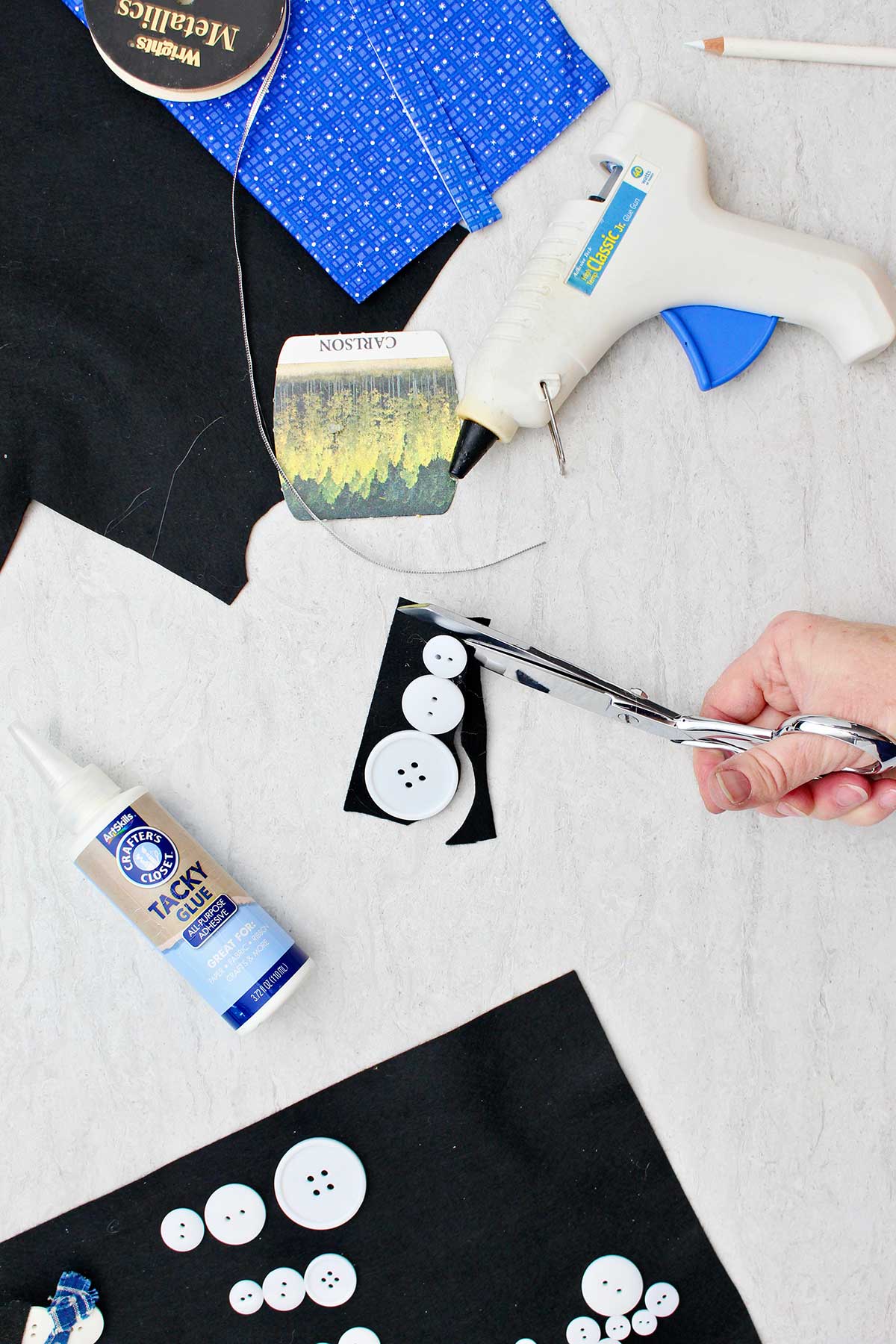 Person cutting excess black felt off the backing of a button snowman with other supplies near by.