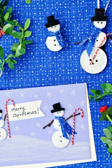 A Christmas card with two button snowmen on it, a snowman ornament and snowman pin made from buttons and fabric surrounded by holly garland against a blue patterned fabric.