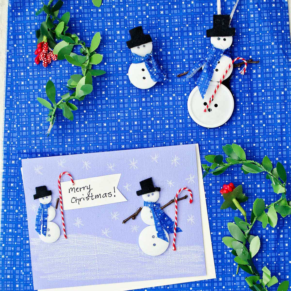 A Christmas card with two button snowmen on it, a snowman ornament and snowman pin made from buttons and fabric surrounded by holly garland against a blue patterned fabric.