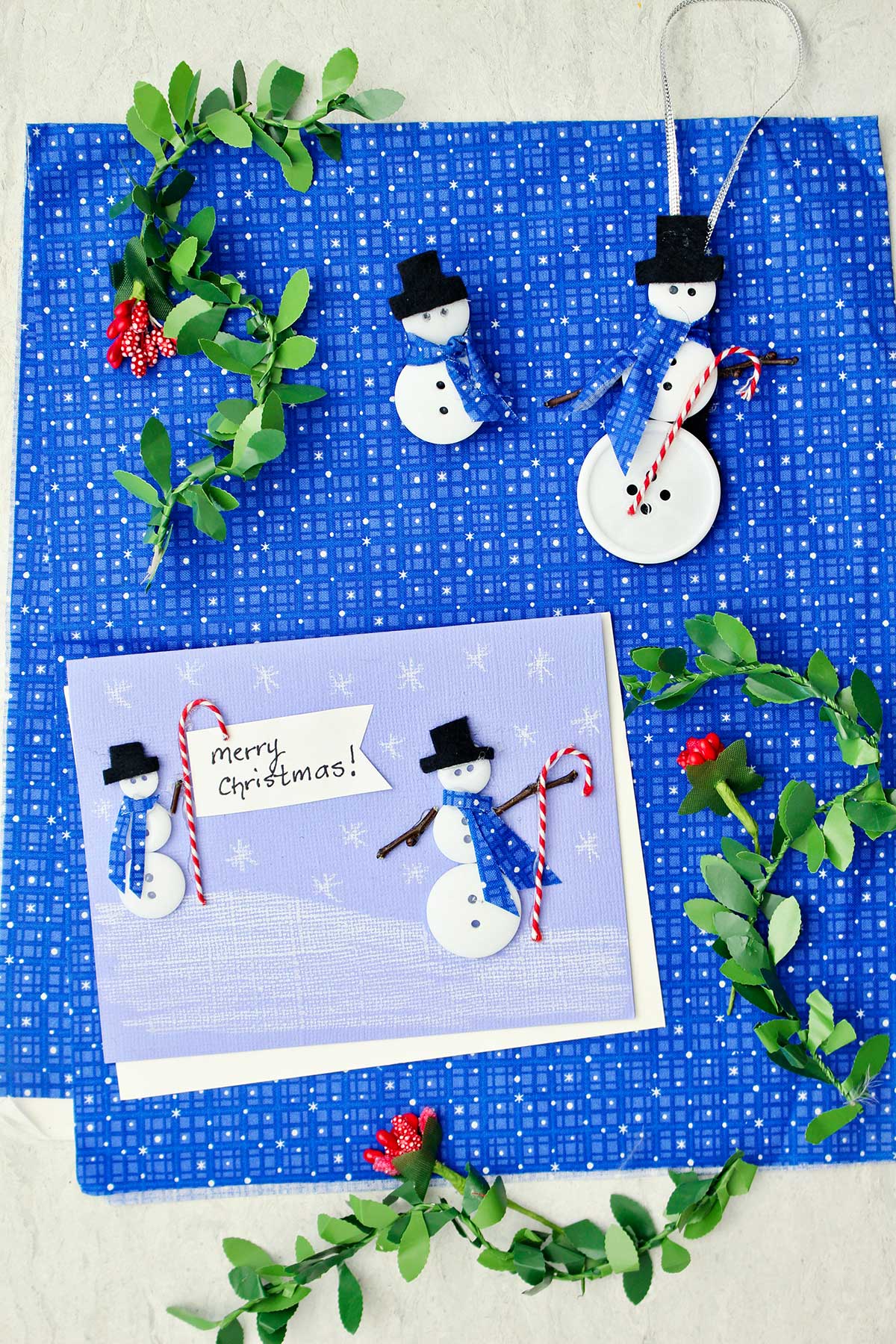 A Christmas card with two button snowmen on it, a snowman ornament and snowman pin made from buttons and fabric surrounded by holly garland against a blue patterned fabric.