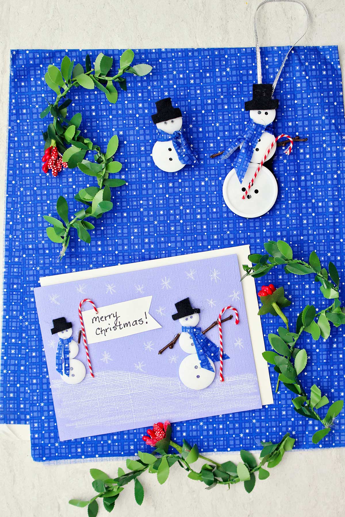 A Christmas card with two button snowmen on it, a snowman ornament and snowman pin made from buttons and fabric surrounded by holly garland against a blue patterned fabric.