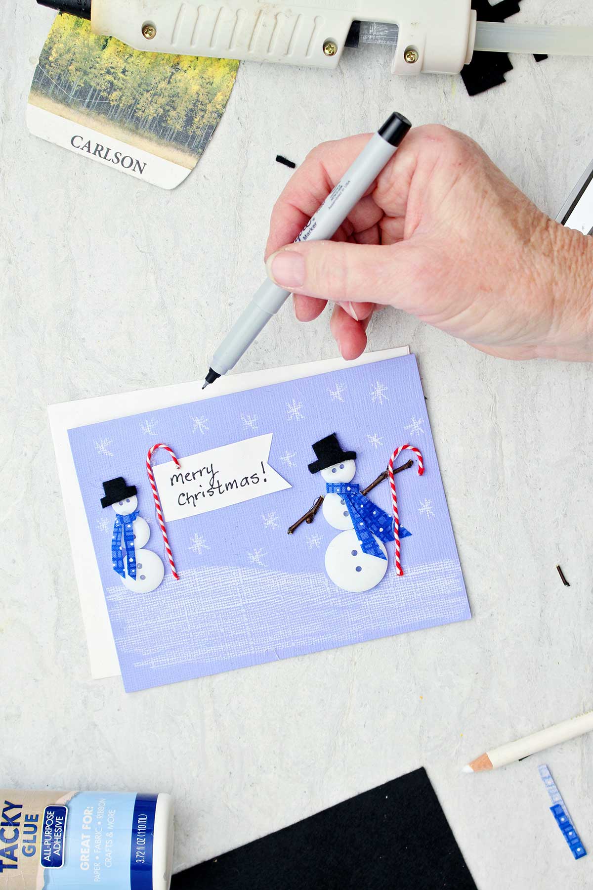 Person adding "Merry Christmas" with black sharpie to a button snowman Christmas Card with other supplies near by.