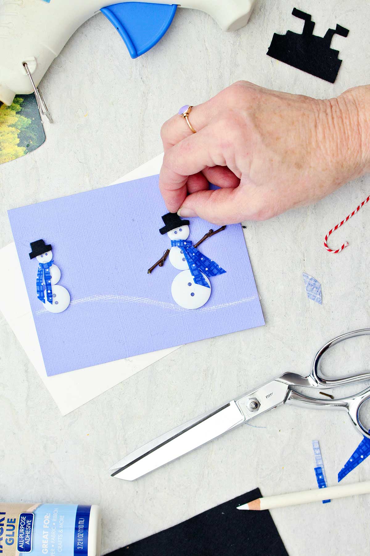 Person adding a hat on a button snowman on a lilac colored Christmas card.