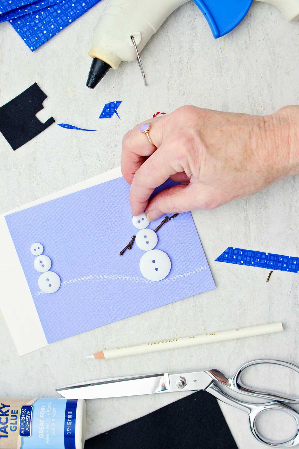 Person adding a button to make the head of a snowman on a lilac colored Christmas card.