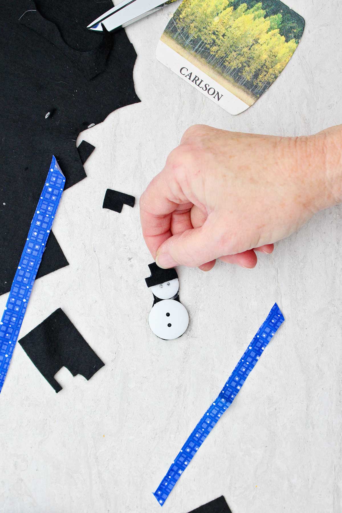 Person adding black top hat to a button snowman with scraps of fabric near by.