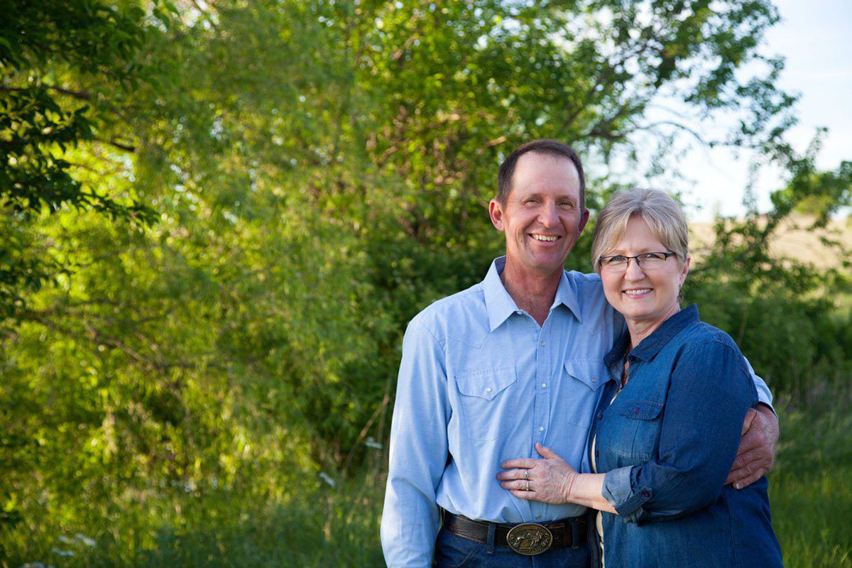 A blonde woman and dark haired man dressed in blue standing in front of some trees.