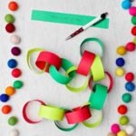 Paper chain made from light green, red, and green construction paper with indoor activities written on them with colorful pom-poms around. Strip of paper saying "Make a pop up card" and pen near by.