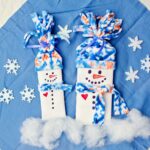 Two completed candy bar snowman stocking stuffers with fleece hats and scarves against a blue cloth with sparkly snowflake decorations.